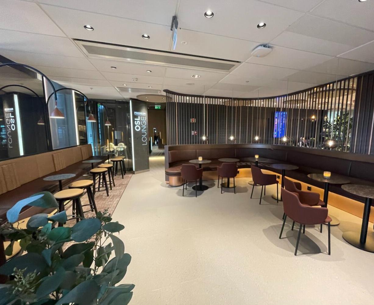 Romslig flyplassloungeinteriør med moderne design, med brune seter, runde bord, og stemningsbelysning, som gir et behagelig venteområde for reisende