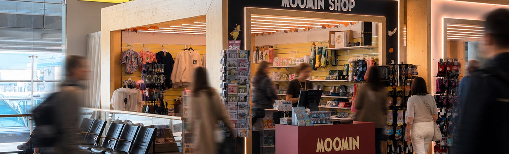 Moomin Shop utsalgskiosk i en flyplassterminal med reisende som går forbi og fargerikt tilbehør utstilt