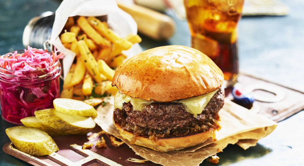 Saftig cheeseburger med sprø pommes frites, sylteagurk og rød coleslaw, ved siden av et forfriskende glass brus
