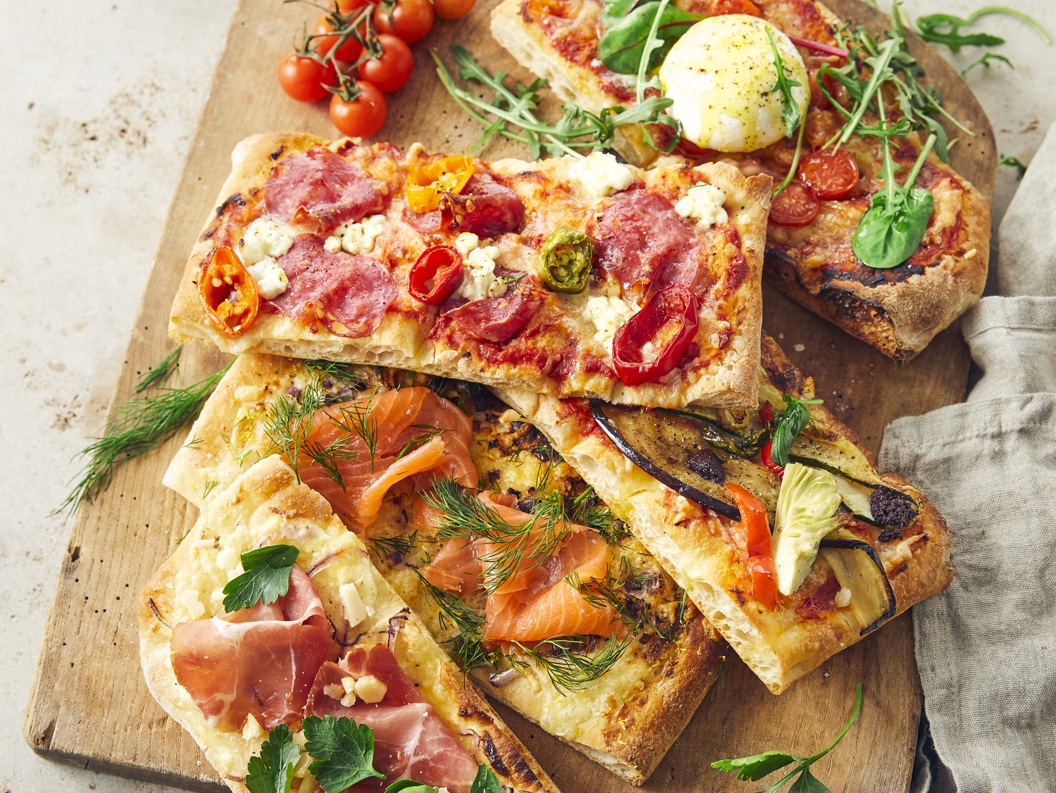 Assorterte gourmetflatbrødpizzaer på et trefat med toppings som inkluderer prosciutto, røkt laks, ruccola og ferske tomater, ledsaget av dippesauser på siden