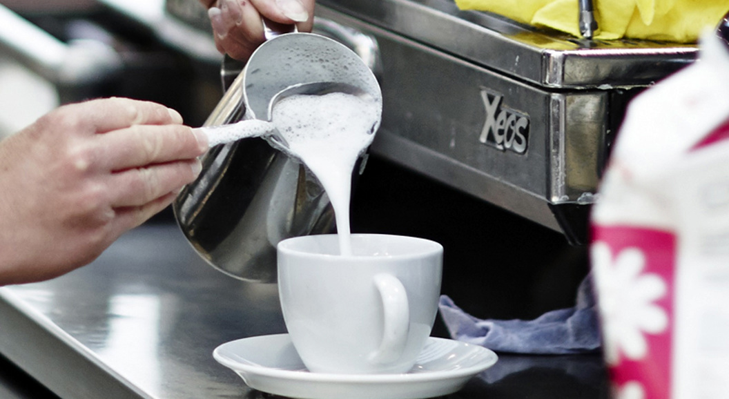 Barista som heller steamet melk i en hvit kaffekopp ved siden av en espressomaskin i en kafé