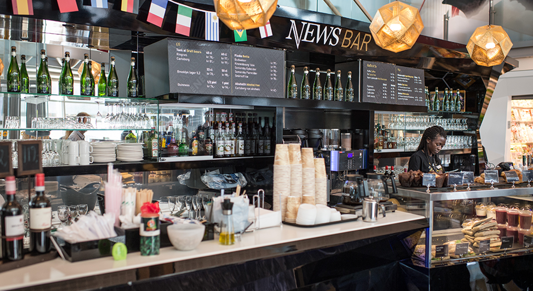 Moderne kafébarinteriør med utvalg av drikkevarer, smørbrød- og bakevaredisplay og en person som serverer bak disken på News Bar, dekorert med internasjonale flagg og varm belysning
