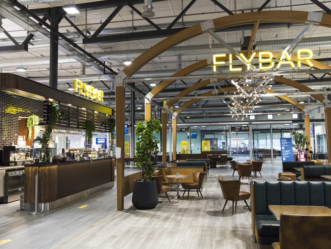 Moderne flyplassbar med stilige sitteplasser og opplyst "FlyBar"-skilt, med buede trebjelker og moderne innredning
