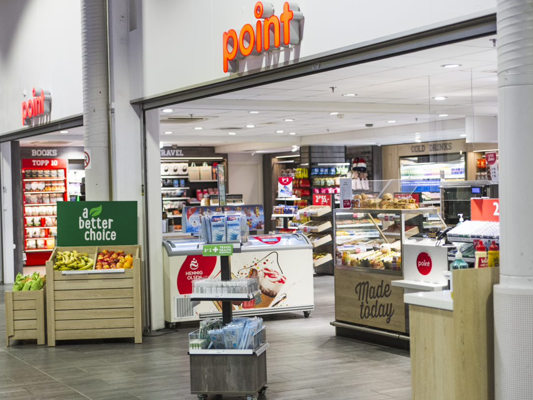 Inngang til en nærbutikk med utstilling av fersk frukt, magasiner, kalde drikker og en delikatesseavdeling med skiltet "Laget i dag". Ulike produkter er tilgjengelige for kunder