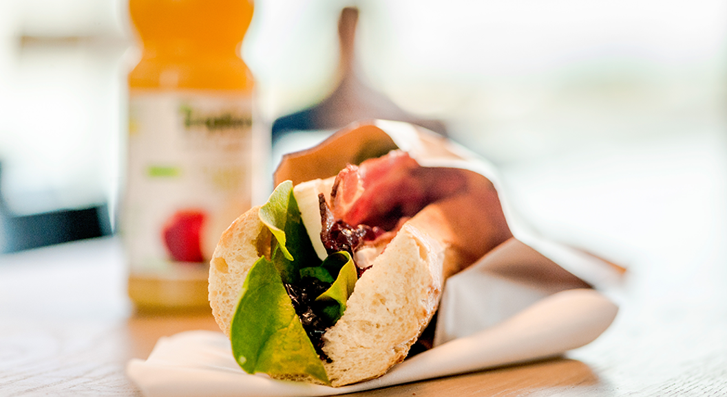 Fersk sandwich med salat, kjøtt og krydder på et bord, innpakket i papir, med en flaske juice i bakgrunnen
