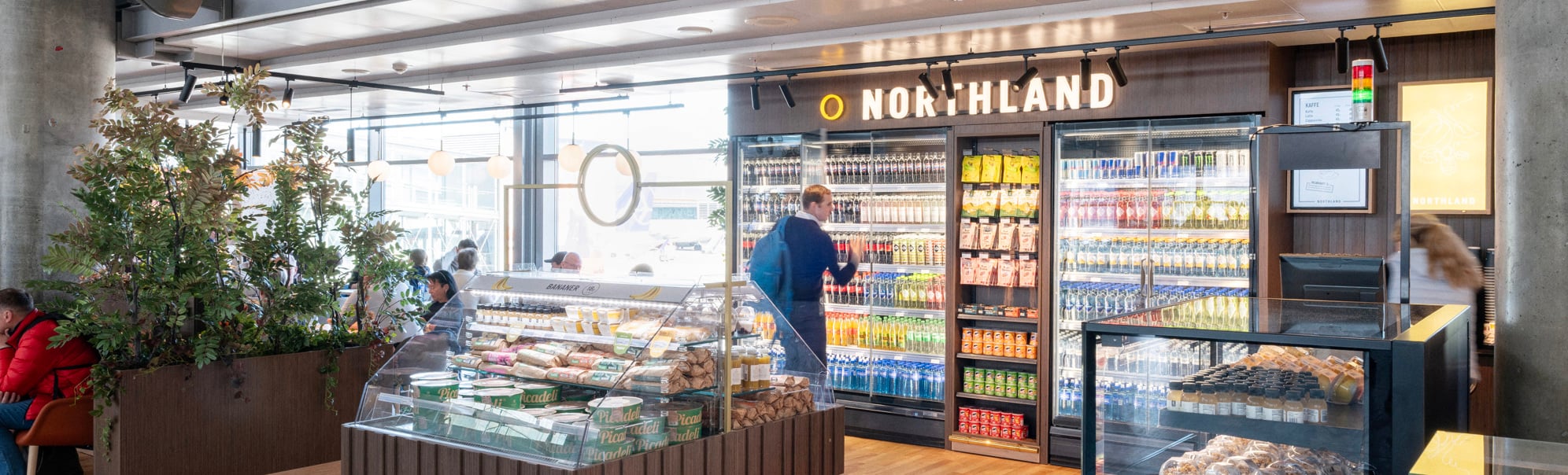 Moderne flyplass-kiosks interiør med kjøledrikker, snacks og fersk matutstilling på Northland-butikk. Kunder utforsker gangene i et godt opplyst, åpent område med tregulv og moderne designelementer