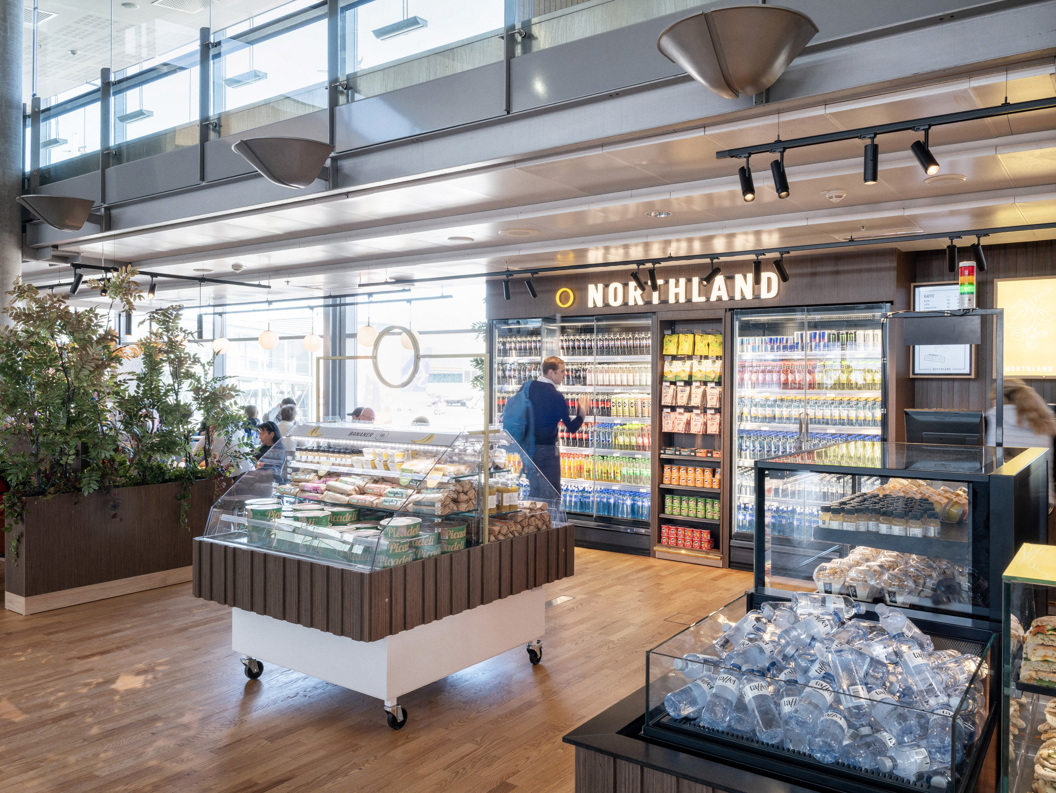 Moderne flyplass-kiosks interiør med kjøledrikker, snacks og fersk matutstilling på Northland-butikk. Kunder utforsker gangene i et godt opplyst, åpent område med tregulv og moderne designelementer