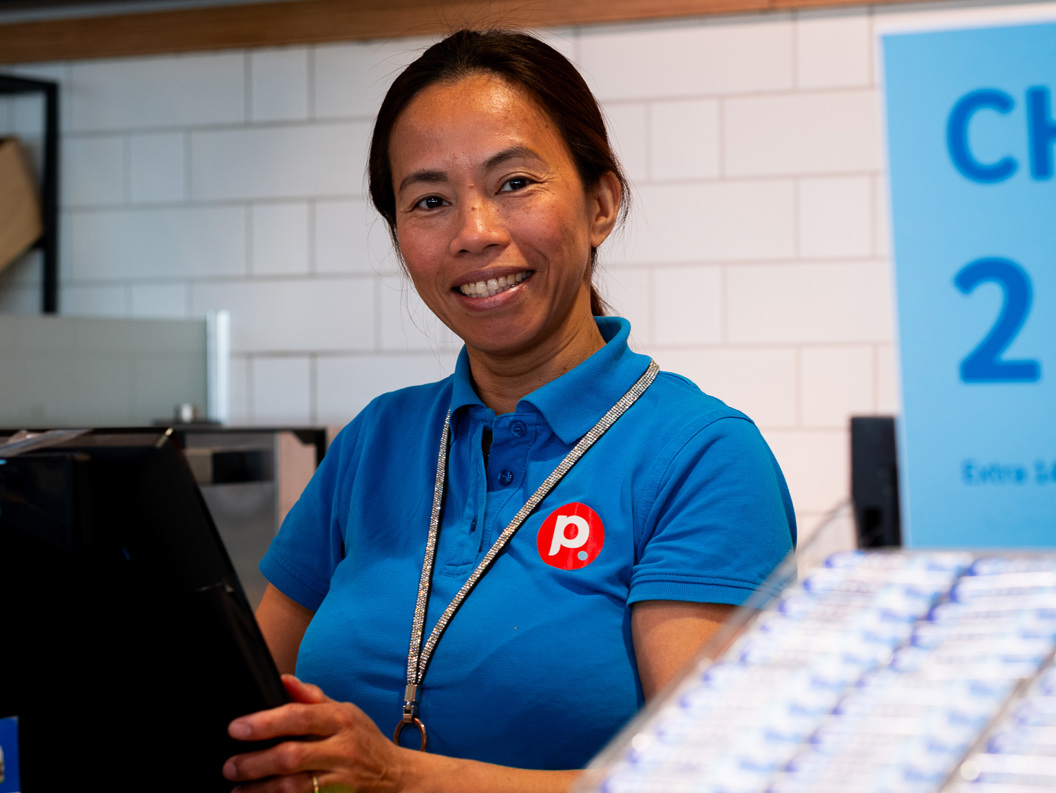 Kvinne i blå uniform jobbar bak kassa i ein butikk, smilande med salgsplakat i bakgrunnen