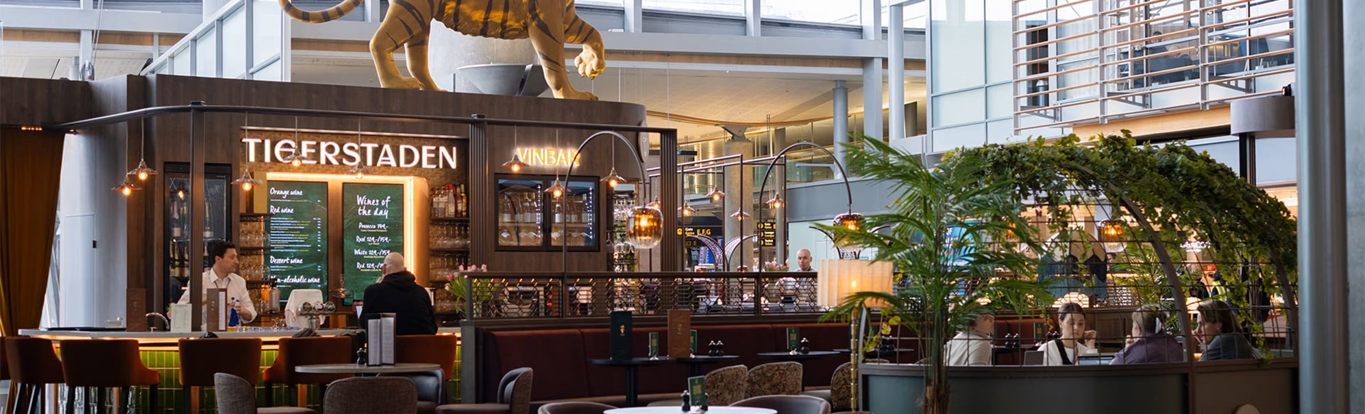 Tigerstadens vinbar på ein flyplass med ein stor tigerfigur på taket. Moderne interiør med sitteplassar og skilt for portar E, F, G i bakgrunnen