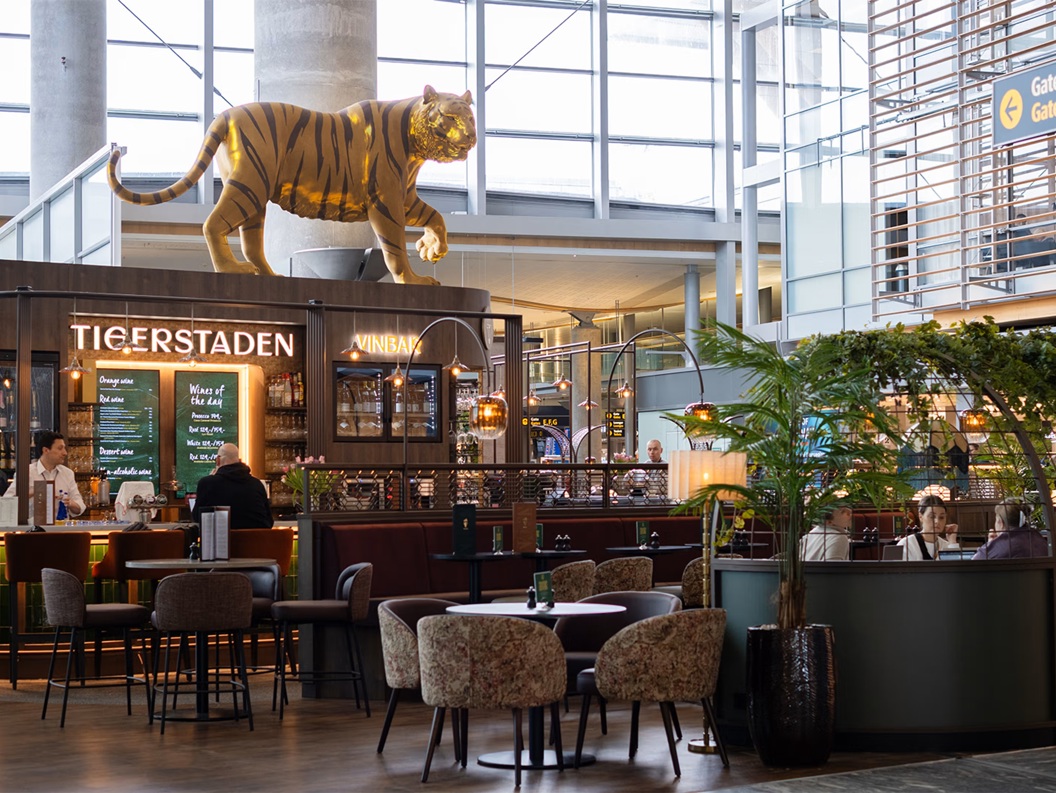Tigerstadens vinbar på ein flyplass med ein stor tigerfigur på taket. Moderne interiør med sitteplassar og skilt for portar E, F, G i bakgrunnen