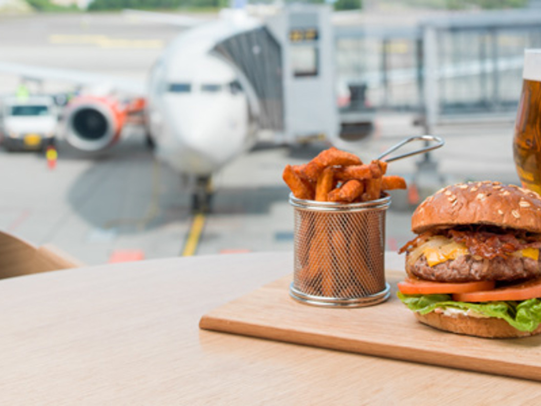 Gourmetburger med pommes frites og et glass øl på et bord med utsikt over en rullebane på flyplassen med et parkert fly i bakgrunnen