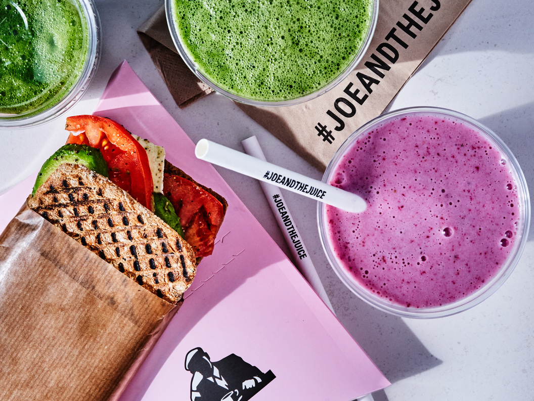 Juice bar-måltid med en grillet sandwich med avokado og tomat, ved siden av grønne og lilla smoothies på et bord, merket med #JoeAndTheJuice