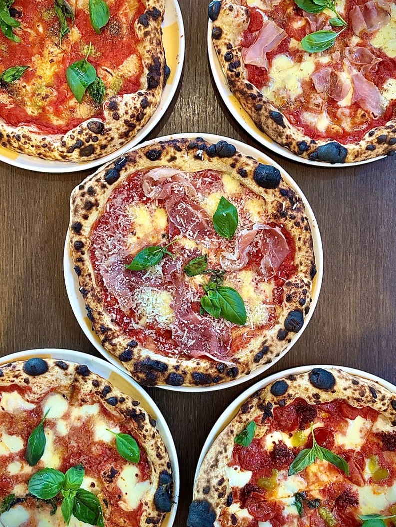 Fem håndverks-pizzaer med ulike toppinger inkludert basilikum, ost og spekemat på et trebord