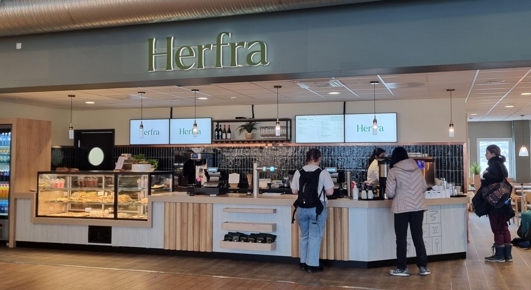 Interior view of a modern cafe named Herfra with customers ordering at the counter. The cafe features a display case with pastries, hanging lights, and digital menu screens above the counter