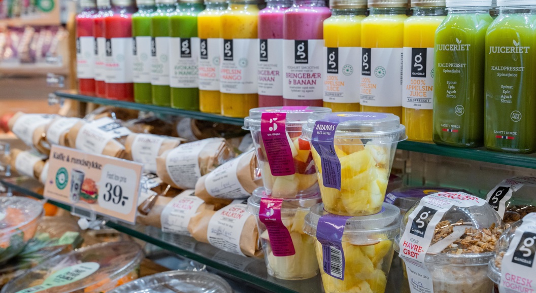 Supermarkedshylle som viser et utvalg av ferske juicer i fargerike flasker, delipakker og snackbeholdere med ananasbiter og granolayoghurt