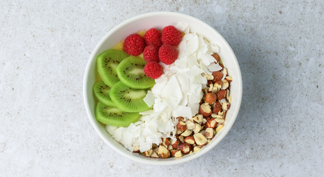 Yoghurtbolle med skiver av kiwi, bringebær, kokosflak og nøtter på ein lys overflate