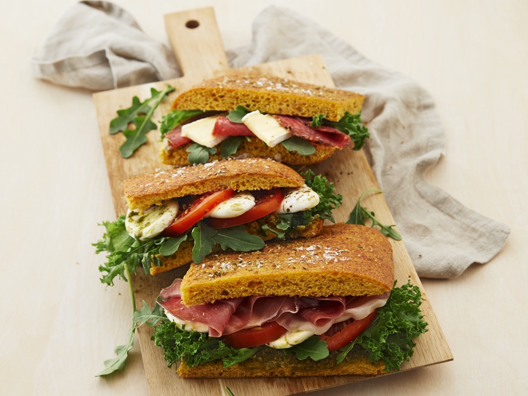 Three gourmet sandwiches with prosciutto, cheese, tomato, and arugula on a wooden board, styled with a linen cloth