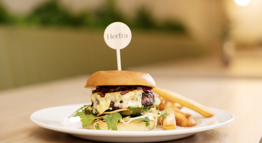 Gourmet cheeseburger på en hvit tallerken med pommes frites, garnert med grønnsaker og toppet med en tannpirker med merkevare, servert på et trebord