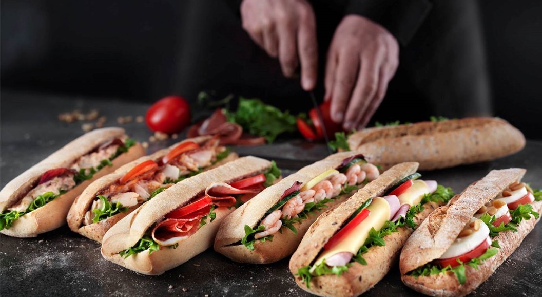 Bilete av ferske baguettar fylt med salat, skinke, reker og ost. Ein person skjer tomatar i bakgrunnen. Mørk flate