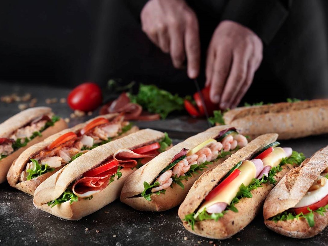 Bilete av ferske baguettar fylt med salat, skinke, reker og ost. Ein person skjer tomatar i bakgrunnen. Mørk flate