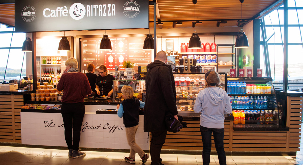 Folk i kø ved Caffè Ritazza på flyplass; kioskseljar lagar bestillingar medan diverse drikkevarer og småretter er utstilte