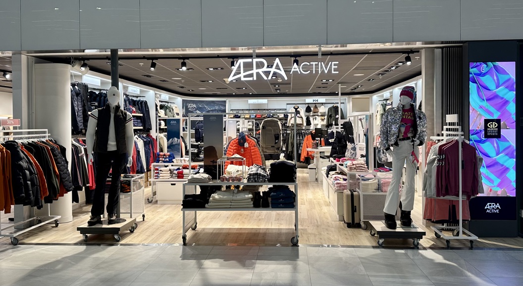 Eksteriør av Aera Active klesbutikk, som viser mannequiner kledd i utendørsklær og fargerike vinterklær, med velorganiserte hyller og stativer som viser forskjellige moteklær inne i det moderne butikklokalet