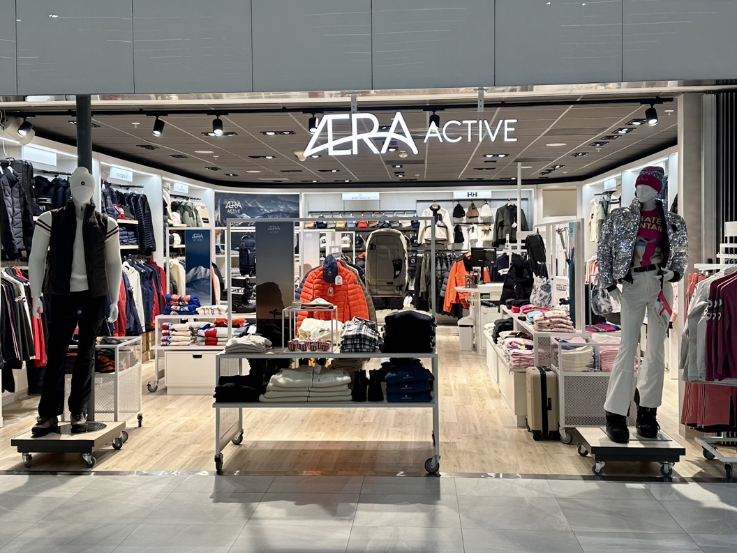 Eksteriør av Aera Active klesbutikk, som viser mannequiner kledd i utendørsklær og fargerike vinterklær, med velorganiserte hyller og stativer som viser forskjellige moteklær inne i det moderne butikklokalet