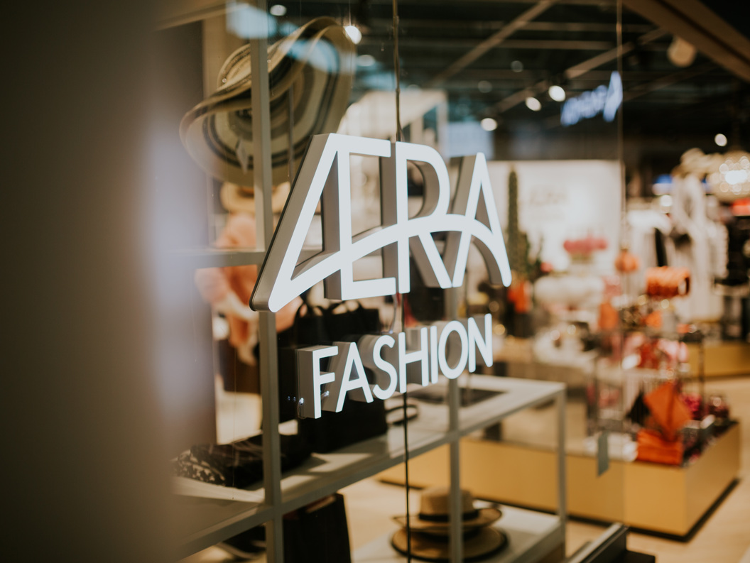 Opplyst "VERA Fashion" skilt på en glassvindu i en moderne motebutikk, som viser frem hatter og stilige klær i et moderne detaljhandelsmiljø