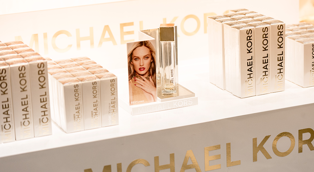 Visning av Michael Kors "Sexy Amber" parfyme med merkede emballasjebokser ryddig arrangert på en butikkhylle