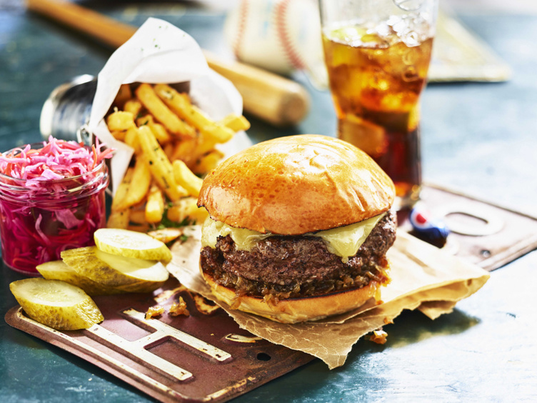 Cheeseburger i burgerbrød med smeltet ost, servert med sprø pommes frites, sylteagurk og en krukke med rød coleslaw, sammen med et glass iskald drikke på et rustikt bord