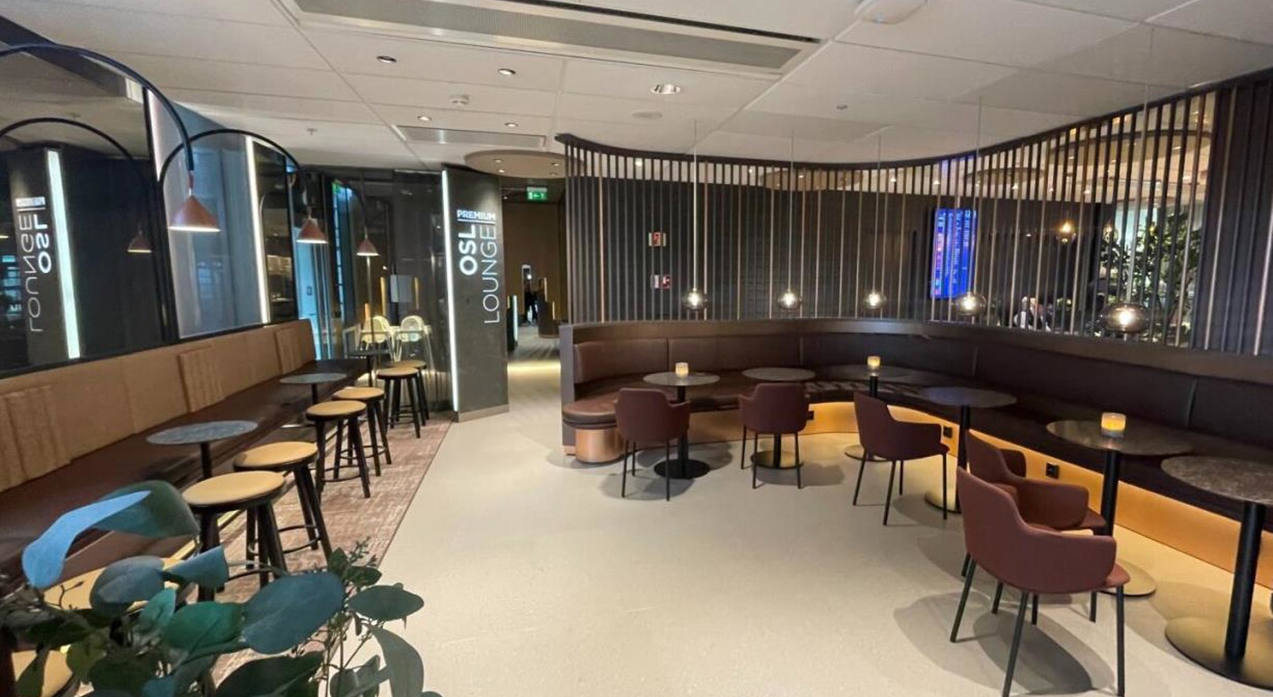 Romslig flyplassloungeinteriør med moderne design, med brune seter, runde bord, og stemningsbelysning, som gir et behagelig venteområde for reisende