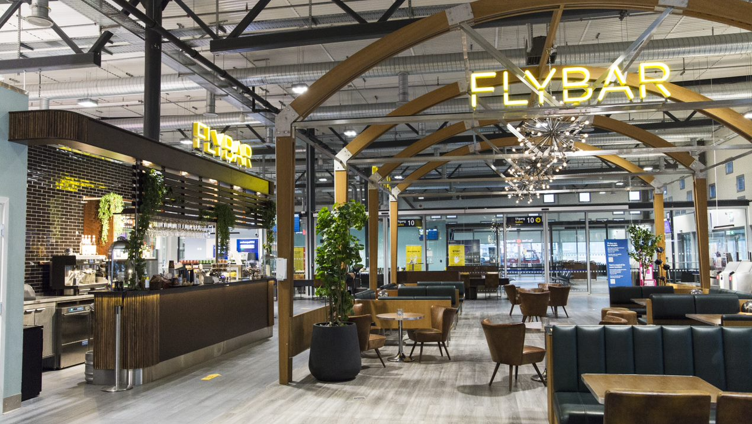 Moderne flyplassbar med stilige sitteplasser og opplyst "FlyBar"-skilt, med buede trebjelker og moderne innredning