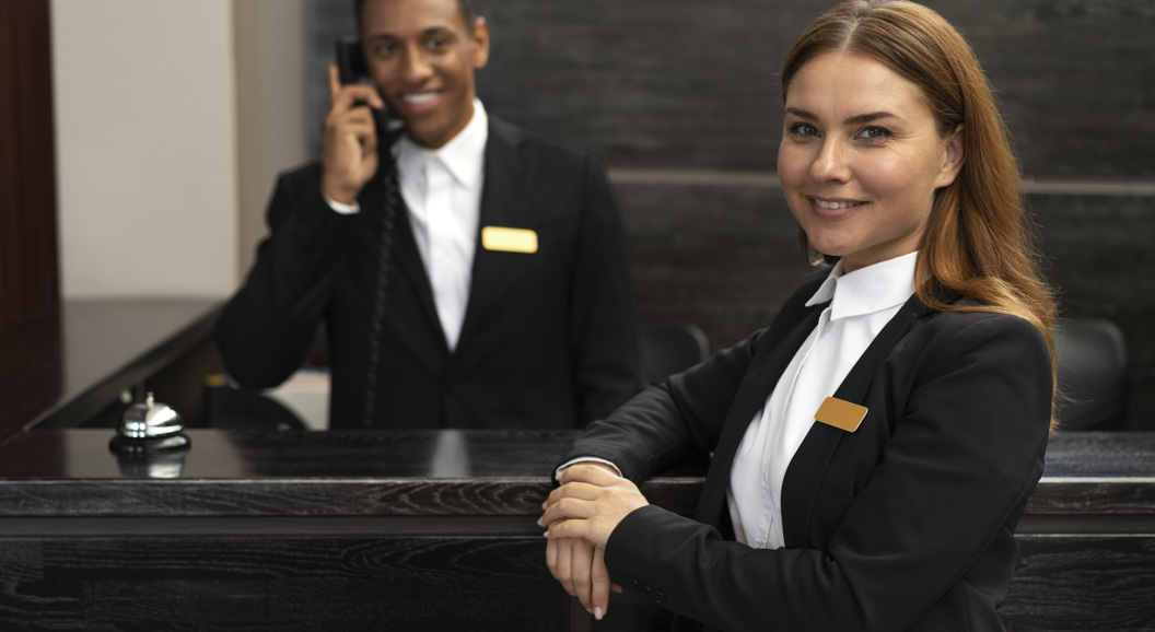 Hotel reception staff smiling, with a man answering the phone and a woman standing in the foreground, both wearing formal uniforms and name badges