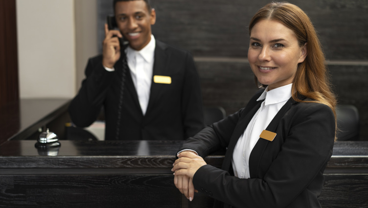Hotellresepsjonspersonale smiler, med en mann som besvarer telefonen og en kvinne som står i forgrunnen, begge iført formelle uniformer og navneskilt