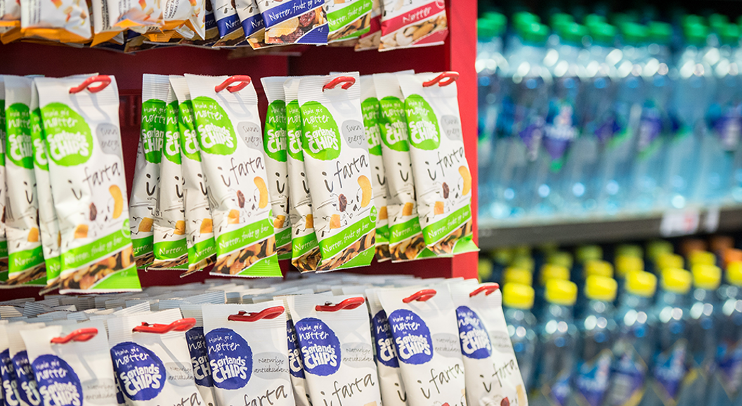 Snack bags on a red retail display shelf, next to a cooler with bottled water in a store setting