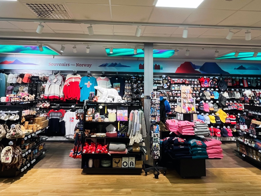 Butikkinteriør av souvenirbutikk i Norge med hyller fulle av klær, leker og suvenirer. Skilt med teksten "Souvenirs - Norway" og neonlogo vises