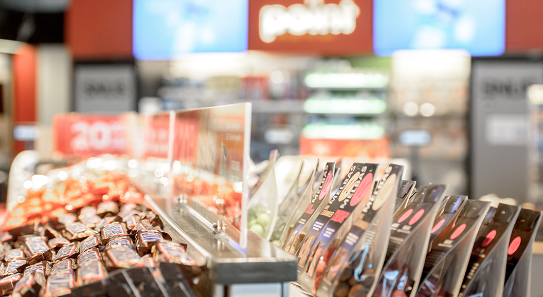 Candy display in a convenience store with chocolate bars and packaged sweets in bright, organized rows. Blurred background features drink coolers and promotional signs