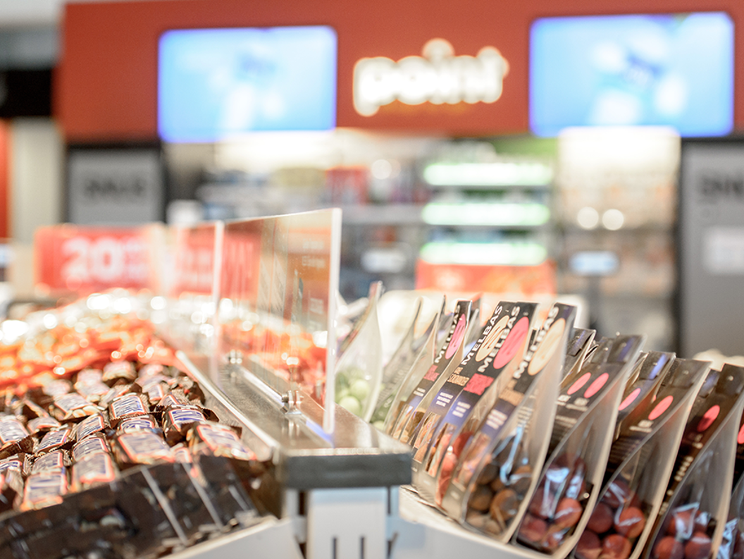 Candy display in a convenience store with chocolate bars and packaged sweets in bright, organized rows. Blurred background features drink coolers and promotional signs