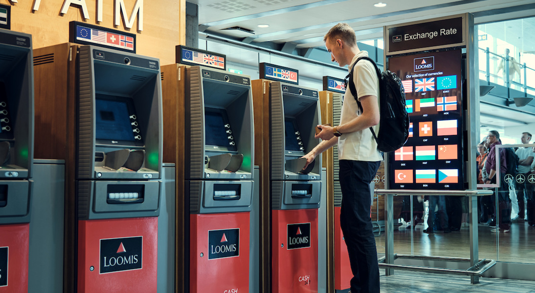 Ein person bruker ein minibank med fleire valutaalternativ ved sidan av ein skjerm som viser valutakursar i ein moderne flyplasshall