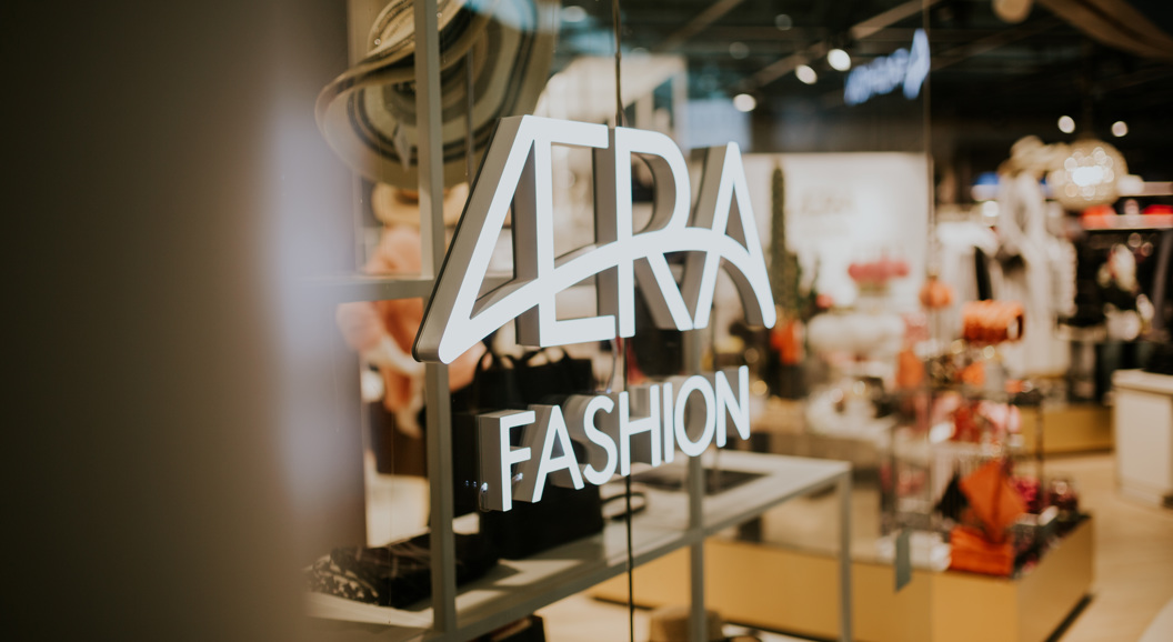 Illuminated "VERA Fashion" sign on a glass window of a trendy fashion store, showcasing hats and stylish clothing in a modern retail environment