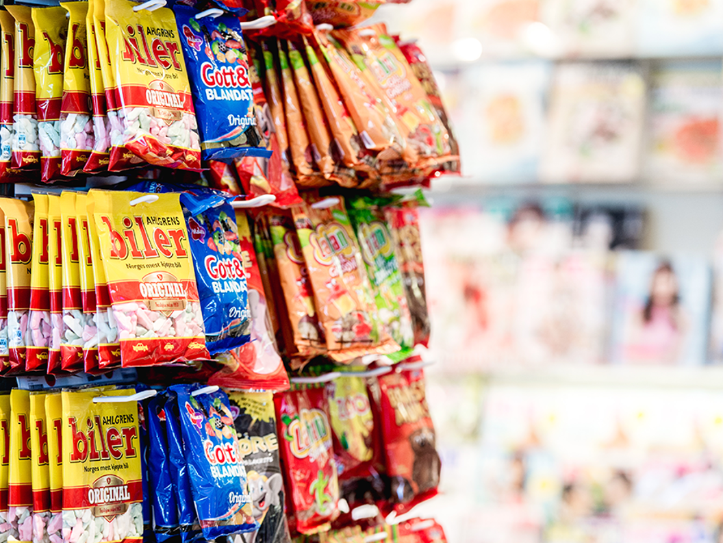 Candy display rack featuring various colorful packaged snacks in a store aisle, with blurred magazine covers visible in the background