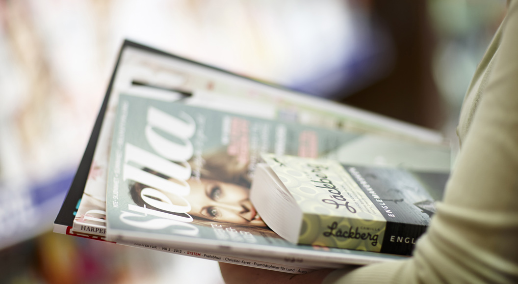 Person holding a stack of magazines and books, featuring a visible "Stella" magazine on top, with a blurred background in a bookstore setting