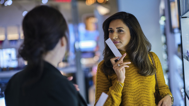 Kvinne i gul genser tester parfyme i en butikk, holder en duftstripe og snakker med en ansatt