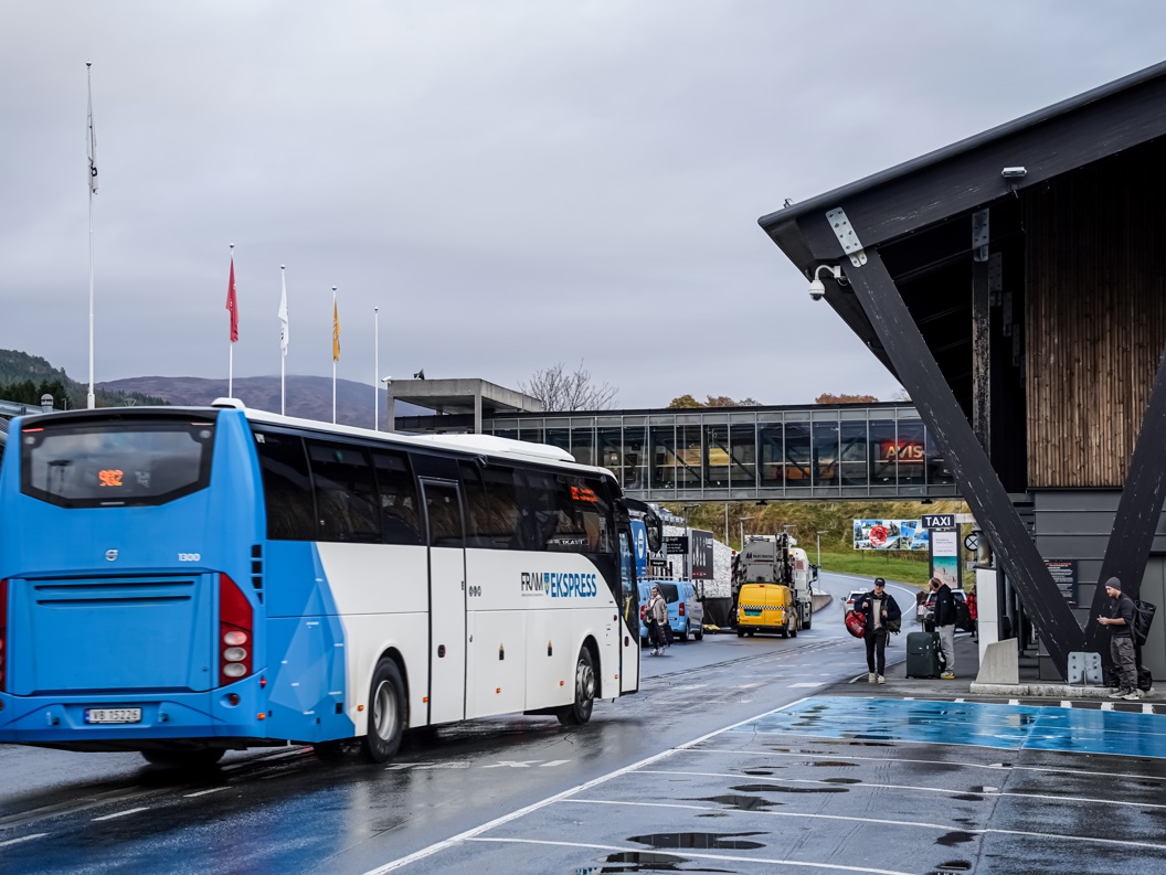 Blå ekspressbuss ved terminal med flere busser og mennesker på en regnfull dag i Norge, omgitt av fjell og moderne arkitektur.