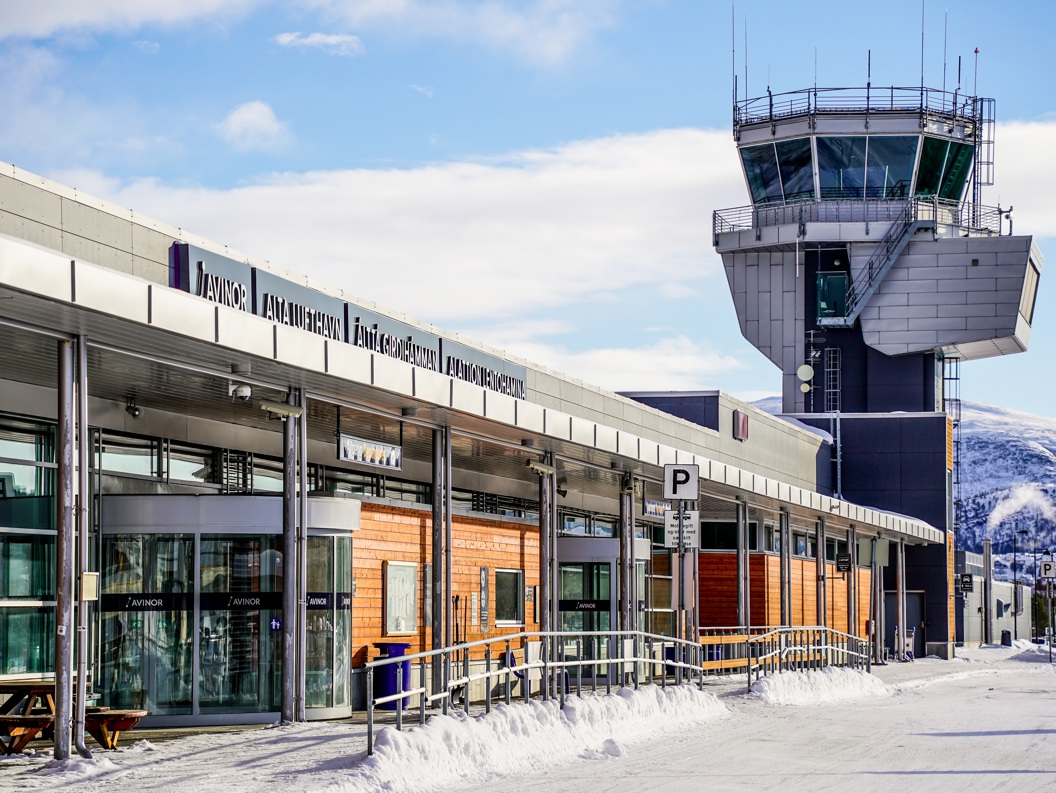 Flyplass med kontrolltårn i vinterlandskap, med skilt for Avinor Alta lufthavn, snødekket bakke og klare blå himmel.