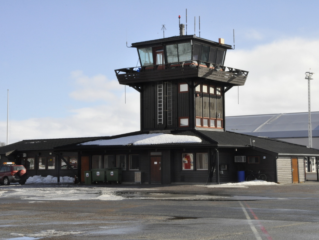 Control tower at Røros Airport with snow on the roof, a parking lot in front, and a car to the left. Winter landscape under a clear sky.