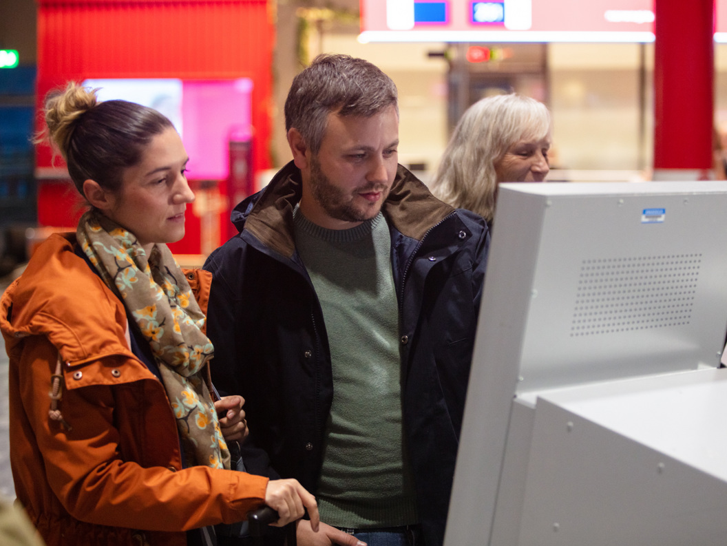 To personer sjekker inn på en digital skjerm på en flyplass. De er kledd i høstjakker.