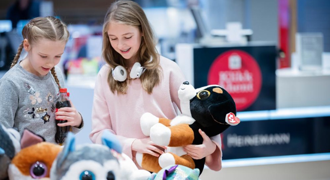 Two children are looking at stuffed animals in a store. One of them is holding a stuffed dog, while the other has a soda bottle.