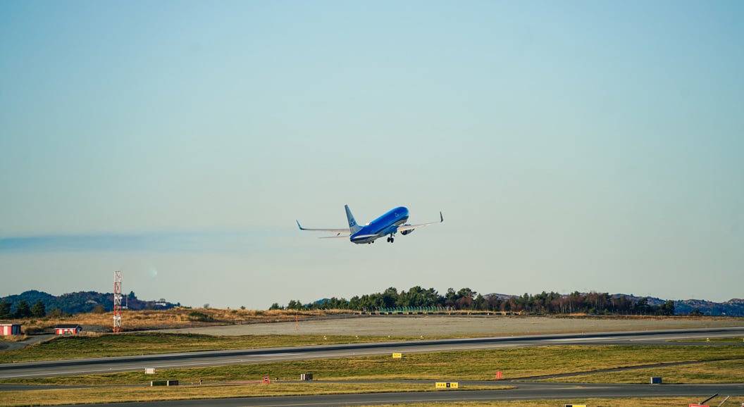 Et blått fly som letter fra en rullebane mot en klar himmel, med åser og trær i bakgrunnen ved en flyplass