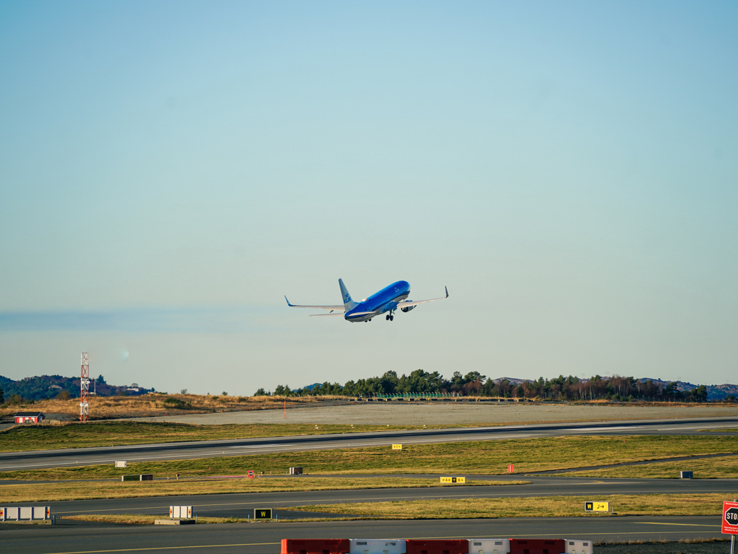Et blått fly tar av fra en rullebane mot en klar himmel, med åser og trær i bakgrunnen på en flyplass